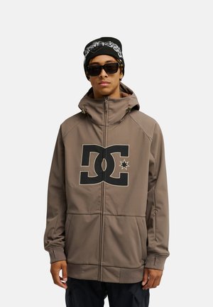 Person mit grauer DC-Kapuzenjacke, schwarzer bedruckter Mütze und dunkler Sonnenbrille steht vor einem schlichten hellen Hintergrund.