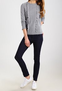 Sudadera gris de punto con patrón texturizado y detalles de volantes, combinada con jeans oscuros y zapatillas blancas. Un atuendo simple y casual.
