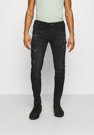 Mann trägt schwarze, abgenutzte Skinny-Jeans mit Farbspritzereien und schwarze Wildlederstiefel, steht vor einem schlichten weißen Hintergrund.