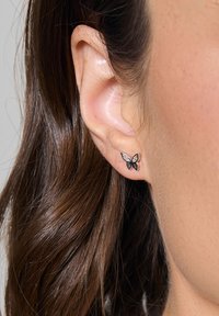 Czarne kolczyki w kształcie motyla z błyszczącym wykończeniem, zdobione przezroczystymi kamieniami wtopionymi w skrzydła. Noszone na uchu modelki, podkreślone brązowymi włosami.