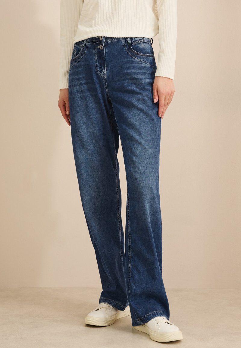 Personne portant un jean bleu à jambes larges, des baskets blanches et un haut blanc à manches longues côtelé, debout à l'intérieur devant un fond uni.