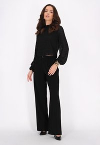 Maglietta nera cropped in tessuto testurizzato con maniche lunghe abbinata a pantaloni neri a vita alta. Gli accessori includono un bracciale dorato.