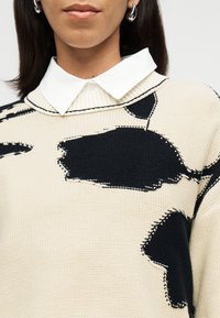 Γυναίκα με λευκό πουκάμισο με γιακά κάτω από ένα εκρού πουλόβερ με μεγάλα μαύρα αφηρημένα σχέδια και ασημένια σκουλαρίκια κρίκους.