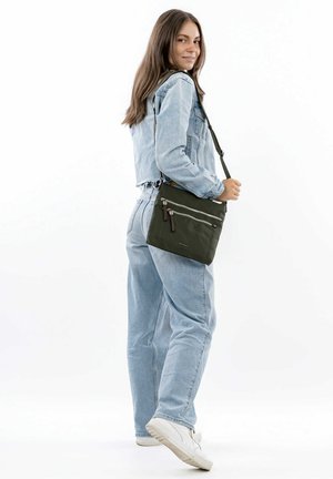Green crossbody bag with two front zippers, featuring a smooth texture and adjustable strap, paired with a denim jacket and jeans.