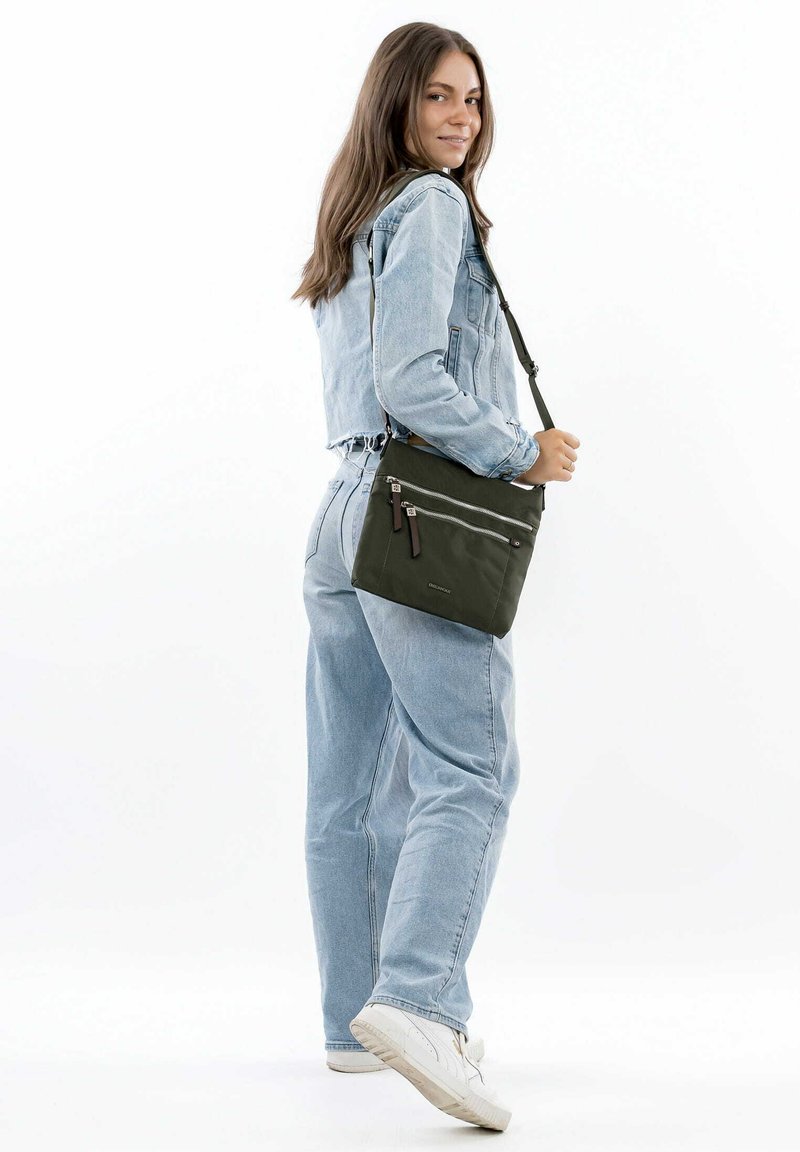 Green crossbody bag with two front zippers, featuring a smooth texture and adjustable strap, paired with a denim jacket and jeans.