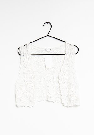 Biały bezrękawnik z koronki o kroju crop top z kwiatowymi wzorami, wiszący na czarnym drucianym wieszaku na białym tle.
