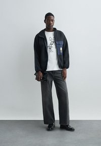 Μαύρο φuzzy μπουφάν με μπλε τσέπη, λευκό μπλουζάκι με γραφικό σχέδιο, φαρδιά μαύρα τζιν και μαύρα παπούτσια. Το μοντέλο κρατά μια μικρή μαύρη τσάντα.