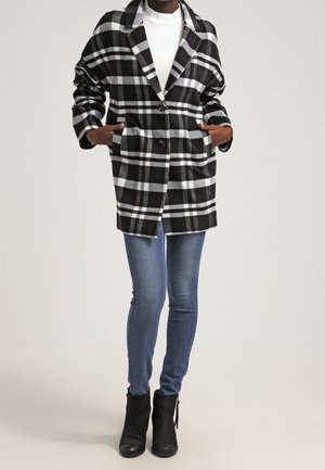 Personne portant un manteau à carreaux noir et blanc sur un top blanc, un jean skinny bleu et des bottines noires, debout les mains dans les poches du manteau.