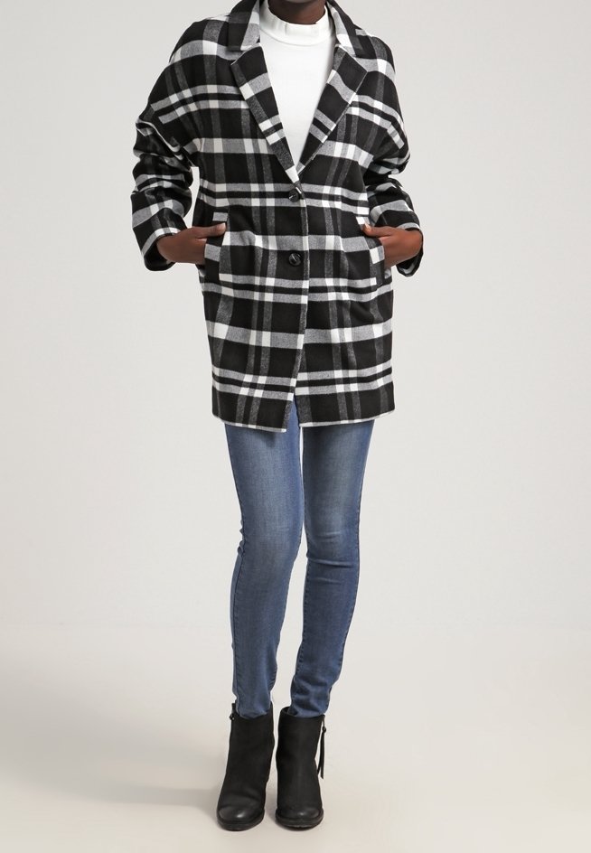 Personne portant un manteau à carreaux noir et blanc sur un top blanc, un jean skinny bleu et des bottines noires, debout les mains dans les poches du manteau.