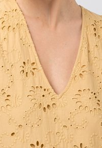 Blusa mostaza con bordado floral, con escote en V y diseños recortados. Tejido suave con un acabado texturizado.