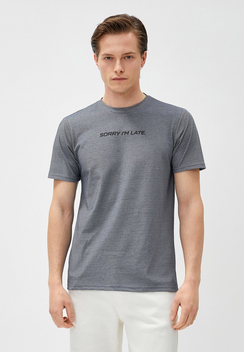 Camiseta gris de manga corta con el texto "LO SIENTO, LLEGO TARDE" en un tono oscuro en el pecho, hecha de una tela suave y texturizada.