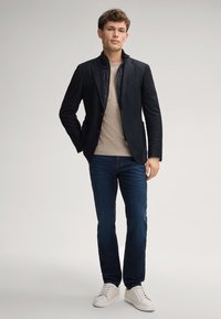 Dunkel maßgeschneiderter Blazer über einem hellen Pullover, kombiniert mit dunkelblauen Jeans und weißen Sneakers. Glatter Stoff mit slim fit und strukturiertem Design.