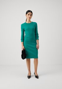 Teal ribbed pitkähiha dress with a fitted silhouette, pyöreä pääntie, ja polven alapuolella pituus, yhdistettynä mustiin kärkitossuihin ja mustaan turkislaukkuun.
