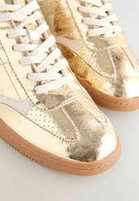 Gold metallic sneakers with a smooth, shiny texture, white laces, perforated detailing, and a brown rubber sole.