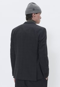 Mand iført en mørkternet jakkesæt, brun turtleneck og grå beanie med "M" mærke, står vendt væk mod en hvid baggrund.