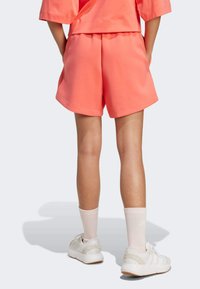 Koralowe szorty o luźnym kroju, teksturowana tkanina, płaski pas, w połączeniu z białymi trampkami i jasnoróżowymi skarpetkami, widok od tyłu.