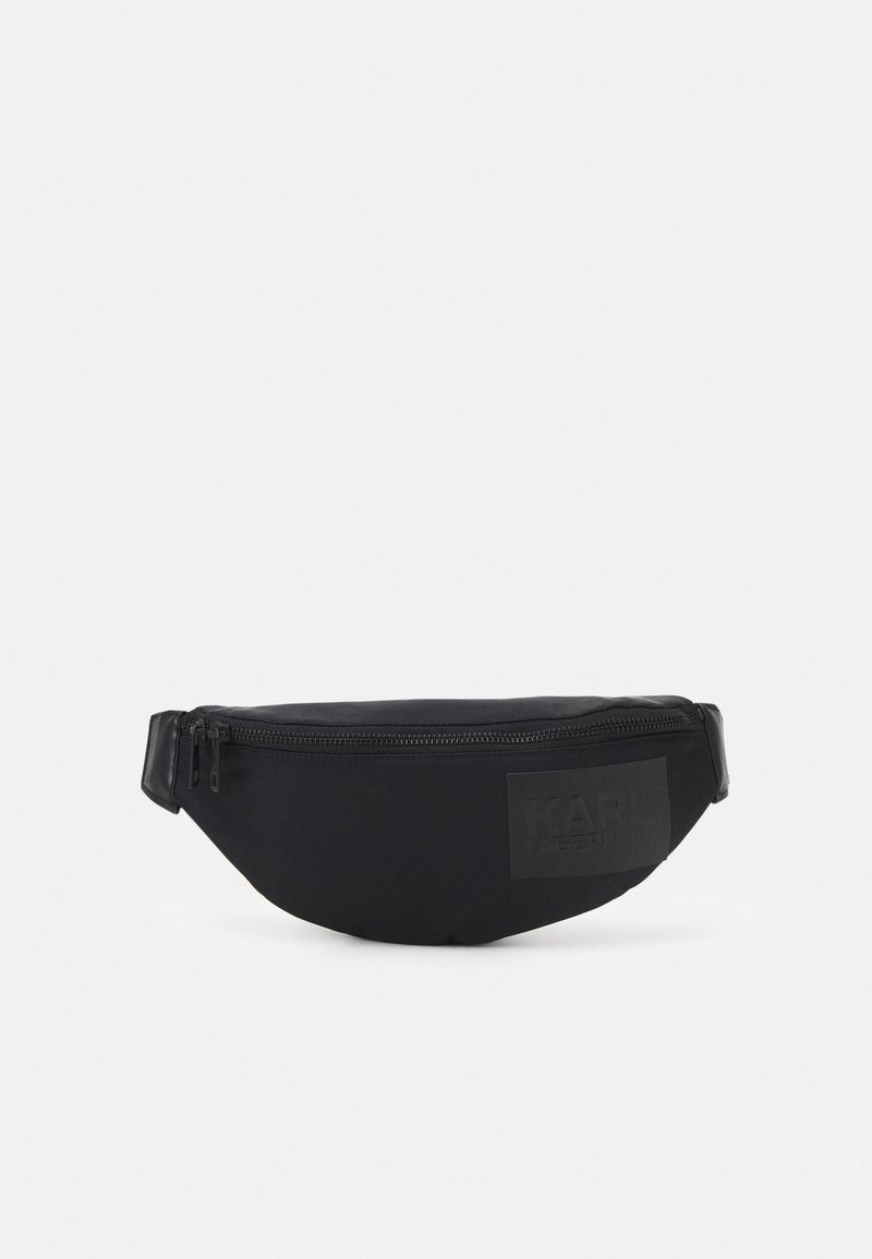 Banane noire zippée avec sangle réglable et logo discret en relief sur le devant, posée sur un fond clair uni.