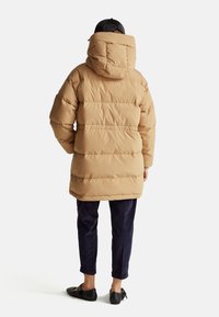 Doudoune marron avec capuche, présentant un design matelassé et une coupe décontractée, assortie à un pantalon bleu marine et des chaussures noires à profil bas.