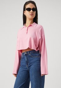Pink cropped long-sleeve polo shirt with a collar and two buttons, paired with high-waisted blue denim jeans.