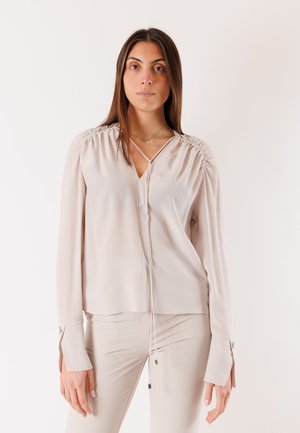 Donna con capelli castani lunghi che indossa una camicetta beige chiaro con spalle arricciate e pantaloni abbinati su uno sfondo bianco uniforme.