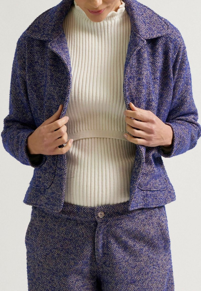 Lila Fischgrätjacke mit Kragen, kombiniert mit einem weißen gerippten Pullover. Die Jacke hat eine lockere Passform und sichtbare Nahtdetails.