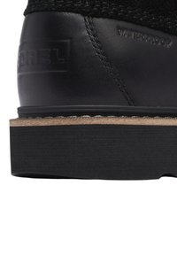 Mustas veekindel saabas, millel on tekstuuriga ülaserv, sile nahast konts ja tugev must küljekoonus looduslike puidu aktsentidega.