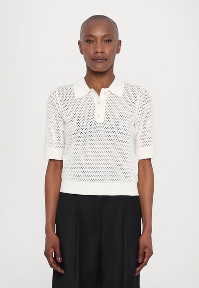 Tricou polo din tricot, de culoare albă crem, cu un model texturat, perforat, mâneci scurte și deschidere cu butoane; purtat cu pantaloni negri largi.