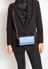 Borsa a tracolla in pelle blu con forma rettangolare, tracolla in catena dorata e chiusura a pattina, indossata su un abbigliamento nero. Texture liscia, design minimale.