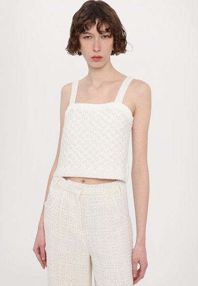 Weißer gestrickter Crop-Top mit quadratischem Ausschnitt und breiten Trägern, mit einem strukturierten Muster. Kombiniert mit hellen, strukturierten High-Waist-Hosen.