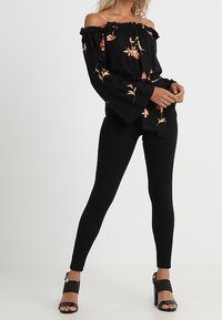 Mujer con blusa negra de flores y manga acampanada, pantalones negros ajustados y sandalias de tacón negro, de pie contra un fondo blanco.