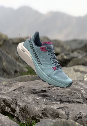 Chaussure de trail Dynafit bleu clair et rose en plein air au-dessus d’un terrain rocheux avec un arrière-plan montagneux flou.