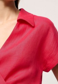 Blusa di lino rossa con colletto a ribattuta e maniche corte, caratterizzata da una texture liscia e da un design a incrocio che crea un scollo a V.