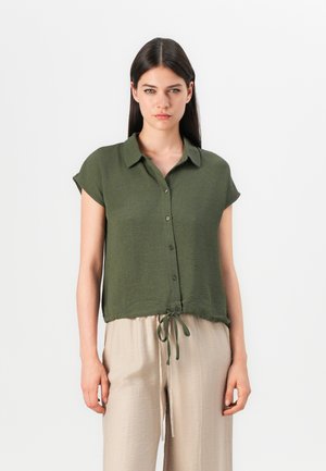 Blusa a maniche corte verde oliva con colletto, bottoni frontali e orlo annodato. Tessuto leggero e strutturato. Abbinata a pantaloni beige dal fit morbido.