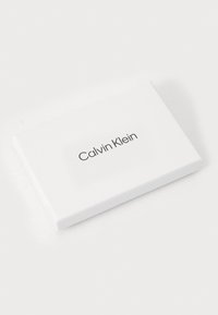 Scatola rettangolare bianca con angoli arrotondati, caratterizzata da un design minimalista e con il logo "Calvin Klein" in rilievo di colore marrone scuro.
