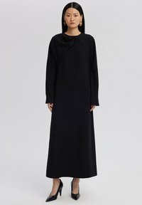 Svart, långärmad klänning i en enfärgade design med rund halsringning och draperad detalj, tillverkad av ett mjukt material. Kombineras med svarta klackar.