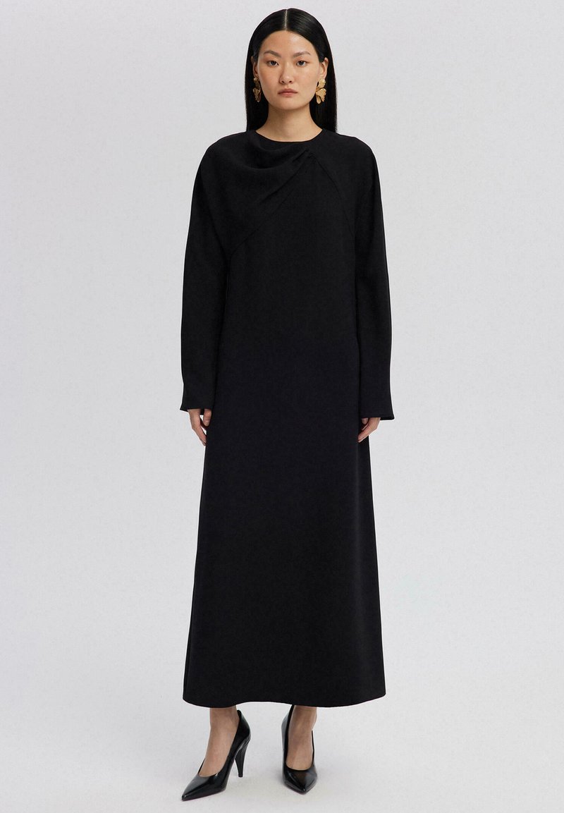 Svart, långärmad klänning i en enfärgade design med rund halsringning och draperad detalj, tillverkad av ett mjukt material. Kombineras med svarta klackar.