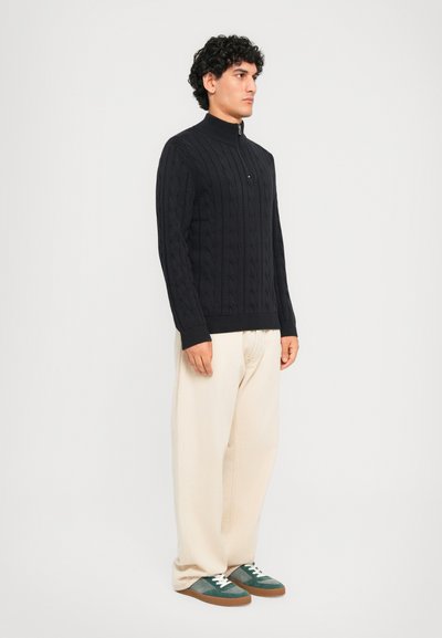 Άνδρας που στέκεται φορώντας ένα μαύρο φούτερ �με πλέξη καλωδίων και φερμουάρ, άνετους μπεζ παντελόνες και πράσινα αθλητικά παπούτσια με λευκές κορδόνες, μπροστά σε άσπρο φόντο.