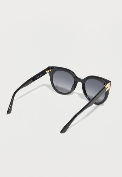 Γυαλιά ηλίου σε σχήμα γάτας, μαύρου χρώματος με φακούς που σκουραίνουν βαθμιαία και χρυσές λεπτομέρειες στα πλαϊνά, απεικονισμένα πάνω σε απλό φωτεινό φόντο.