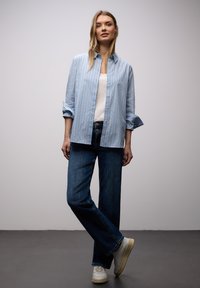 Femme debout, jambes croisées, portant une chemise boutonnée à rayures bleues par-dessus un haut blanc, un jean bleu foncé et des baskets blanches.