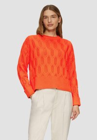 Leuchtend orangefarbener Strickpullover mit einem strukturierten Rautenmuster sowie gerippten Bündchen und Saum, kombiniert mit hellen Hosen.
