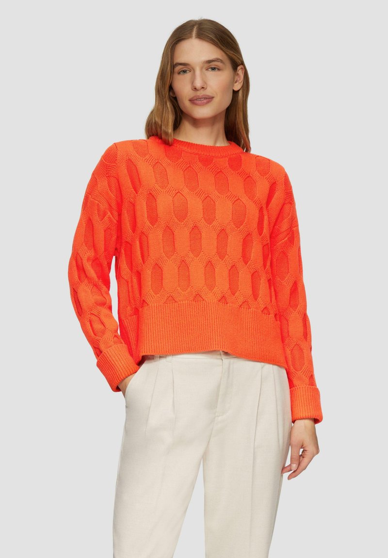 Leuchtend orangefarbener Strickpullover mit einem strukturierten Rautenmuster sowie gerippten Bündchen und Saum, kombiniert mit hellen Hosen.