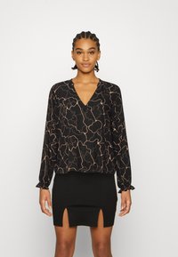 Blusa preta de manga longa com decote em V e padrão abstrato em linhas douradas, combinada com uma saia preta com duas fendas frontais.