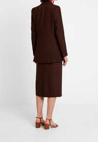 Hnědý barevně sladěný blazer a sukně, se rozparkem na sukni a sandály na blokovém podpatku s kotníkovým páskem. Hladká tkanina se strukturovaným designem.