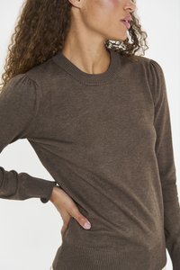Femme aux cheveux bouclés portant un pull en tricot marron à manches longues, se tenant avec une main sur la hanche, tournée vers la droite.
