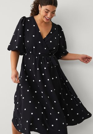 Smilende kvinde, der snurrer rundt i en sort slå-om-kjole med hvide prikker og puffede korte ærmer mod en ensartet lys baggrund.