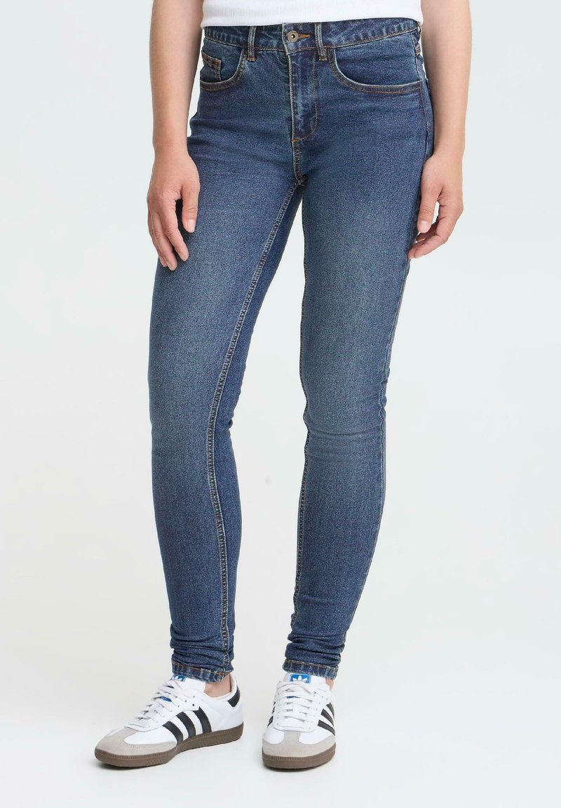 En person som bär slim-fit blå jeans och vita sneakers med svarta ränder, står mot en neutral ljus bakgrund.