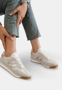 Chaussures de sport beiges avec un dessus en daim et en toile, dotées de trois bandes métalliques, d'une semelle blanche et d'une semelle extérieure en caoutchouc texturé.