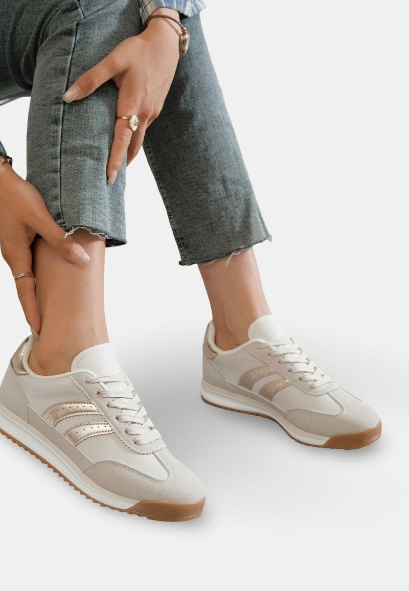 Chaussures de sport beiges avec un dessus en daim et en toile, dotées de trois bandes métalliques, d'une semelle blanche et d'une semelle extérieure en caoutchouc texturé.