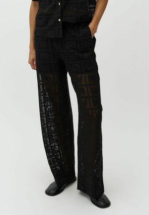 Person iført sorte, gennemsigtige bukser med brede ben og et tekstureret ternet mønster samt sorte sko, stående med den ene hånd i lommen.