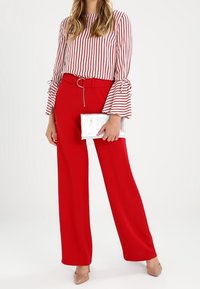 Femme portant un pantalon large rouge avec une ceinture assortie, un chemisier blanc à rayures verticales rouges et manches évasées, tenant une pochette argentée.